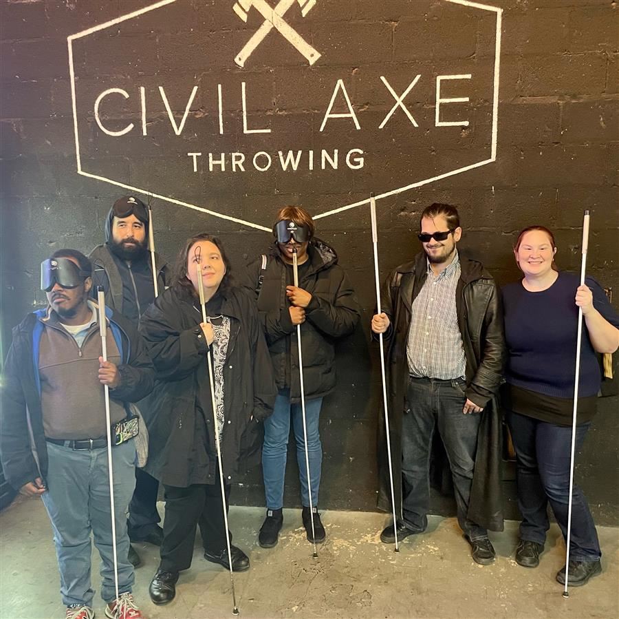  Six people smile to camera in front of a wall with the "Civil Axe Throwing" holding white canes.